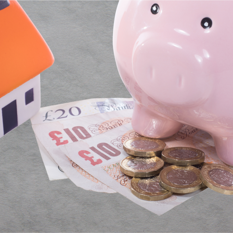 Piggy bank sitting on top of currency next to a miniture house on a grey background.