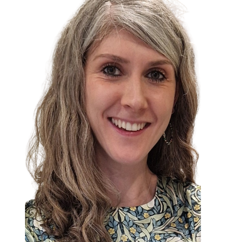 Picture of a smiling woman looking at the camera. She has shoulder length wavy hair is wearing a patterned top