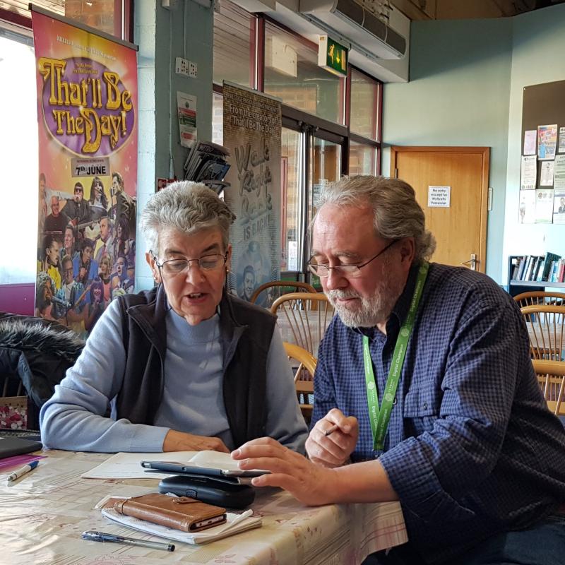 A man and a woman sitting at a table. The man is a digital champion and he is holding a tablet in his hand. He is showing the woman how to use it