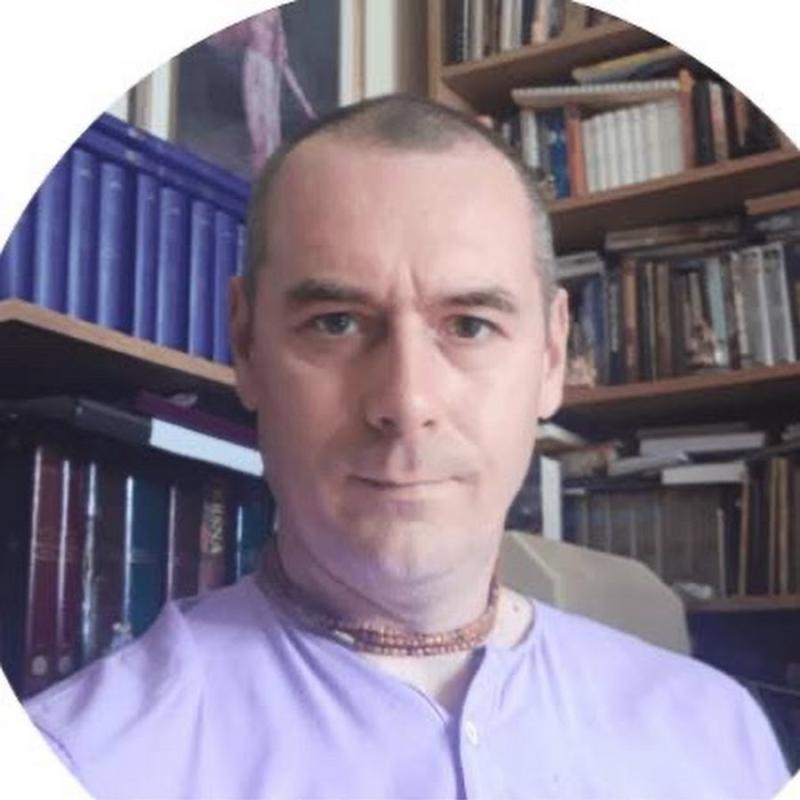 A picture of a man. He is looking at the camera and wearing a light purple shirt. He has beads round his neck.