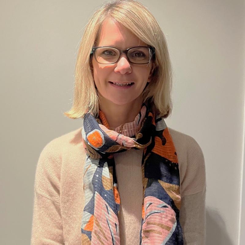 Photo of Aimee Venner. She is smiling at the camera. She has a colourful scarf round her neck, has short blonde hair and is wearing glasses.