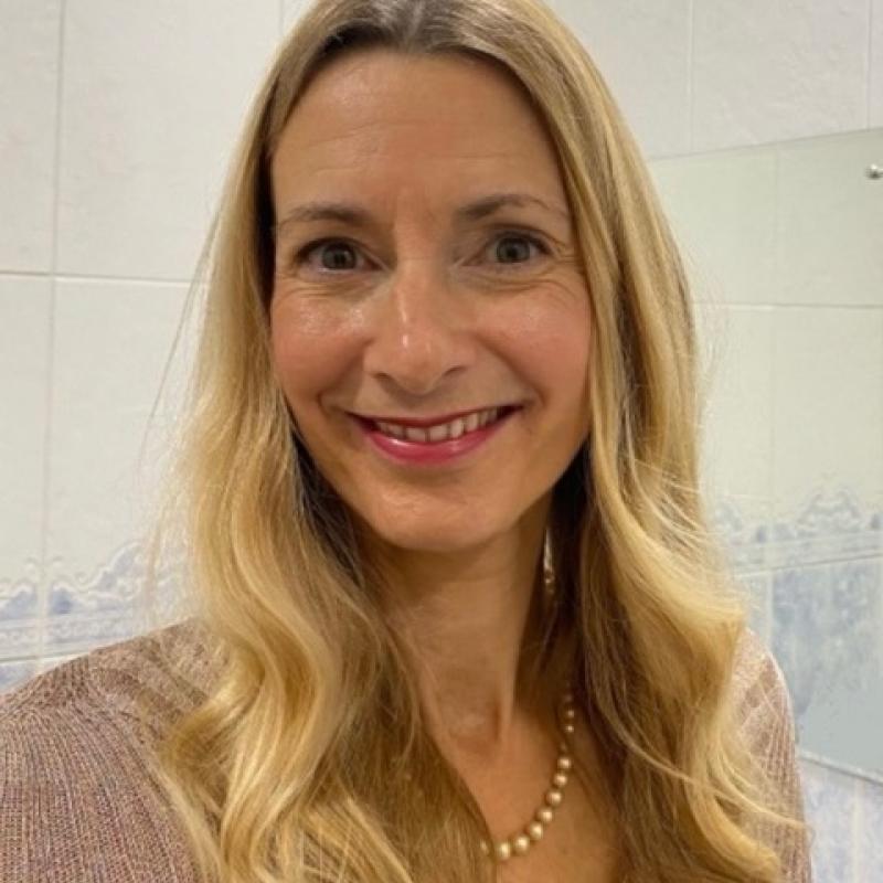 Woman smiling at the camera. She has long wavy blonde hair and has a string of beads round her neck.