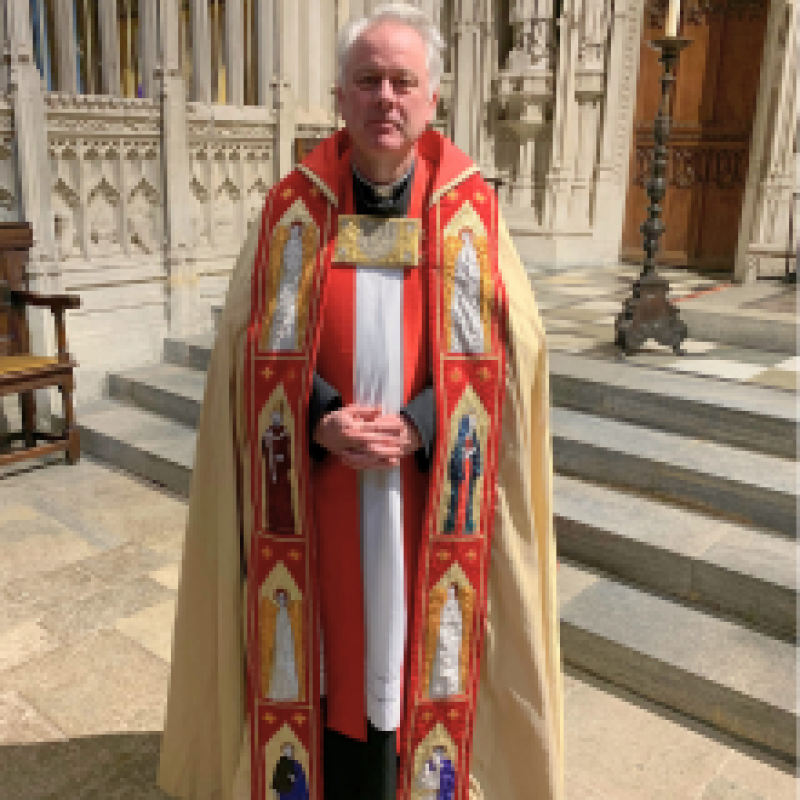 The Revd Will Gibbs. He is standing in front of a cathedral in full robes of red and gold