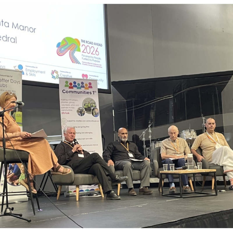 A woman sits on a stool as she facilitates a panel discussion. There are four men sitting in a row in chairs on a stage